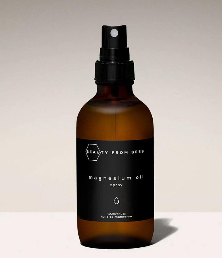 A dark amber glass bottle of "BEAUTY FROM BEES" magnesium oil spray stands on a light surface against a gradient beige background. The bottle has a black label with white text, featuring the brand name at the top and "magnesium oil spray" prominently displayed. A simple droplet icon is also on the label, and the bottle has a black spray nozzle.