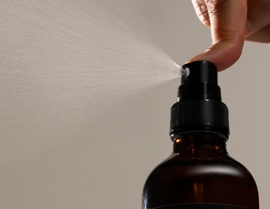 A close-up shows a finger pressing the nozzle of a black spray top on a dark amber glass bottle, releasing a fine mist into the air. The background is a blurred light beige. The bottle's label is partially visible at the bottom.