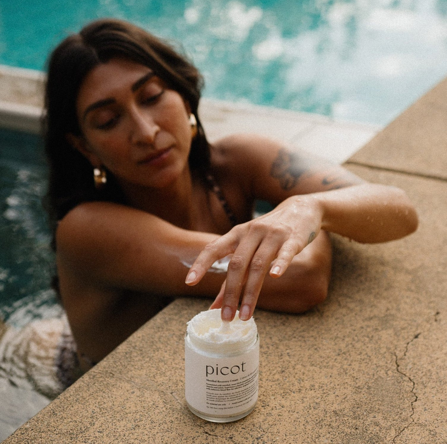 A woman in a bikini is sitting in a pool, reaching toward a jar of "picot Menthol Recovery Cream" that is placed on the concrete edge.