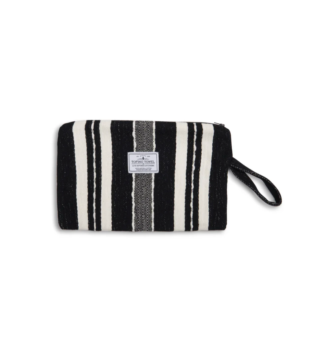 A front-facing flat lay photograph of the closed Mercer Wet-Dry bag. The woven Turkish Cotton exterior features thick, vertical black and ecru (off-white) stripes, with a contrasting black and grey textured panel running down the center. A small "Tofino Towel Co." logo is stitched onto the center panel, and a black woven wrist strap is attached to the upper right corner. The bag rests against a clean, white background.