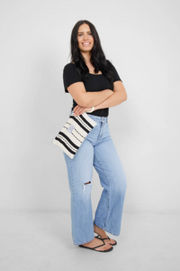 Thumbnail for A smiling woman with long dark hair stands with her arms crossed, holding the ecru and black striped Turkish cotton bag. She is wearing a black top, ripped blue jeans, and black flip-flops.