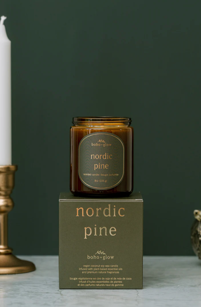 
A close-up photograph of an amber jar candle labeled "nordic pine" by Boho + Glow, sitting centered atop its matching dark olive green box. A brass candle holder and white taper candle are partially visible on the left against a deep green background.