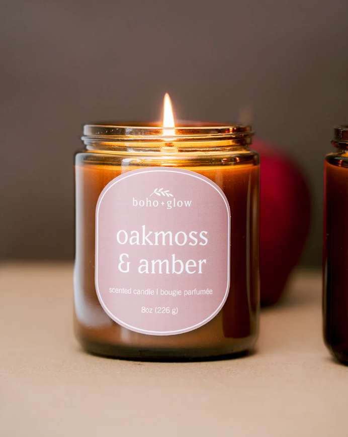 A "boho+glow" scented candle is shown in a glass jar, its wick lit and glowing brightly. The label, with rounded corners, reads "oakmoss & amber."