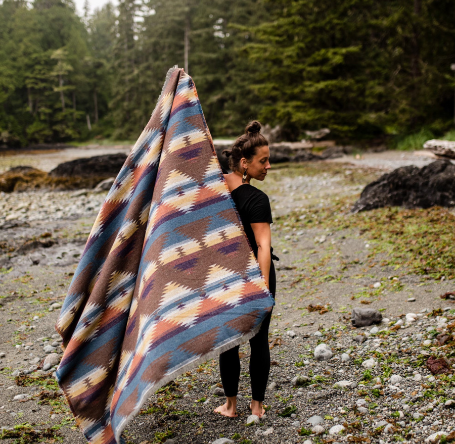 
A woman with her hair in a bun, wearing a black top and black leggings, stands barefoot on a rocky beach. She is holding up a large, patterned blanket in shades of brown, blue, orange, and tan, draped over her shoulder. The blanket features a Southwestern-style geometric pattern. In the background, there's a body of water and a lush green forest under an overcast sky.