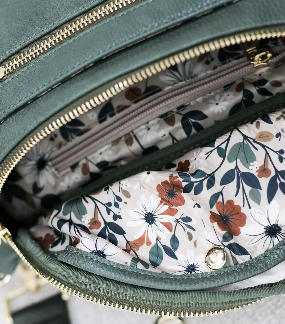 A close-up shot looks down into an open green belt bag. The interior lining is a light cream color with a floral pattern featuring rust-colored, white, and navy blue flowers, along with sage green leaves. A smaller zippered compartment is visible within the main section. The green exterior of the bag has gold zippers.