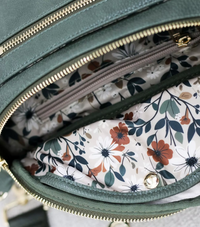 Thumbnail for A close-up shot looks down into an open green belt bag. The interior lining is a light cream color with a floral pattern featuring rust-colored, white, and navy blue flowers, along with sage green leaves. A smaller zippered compartment is visible within the main section. The green exterior of the bag has gold zippers.