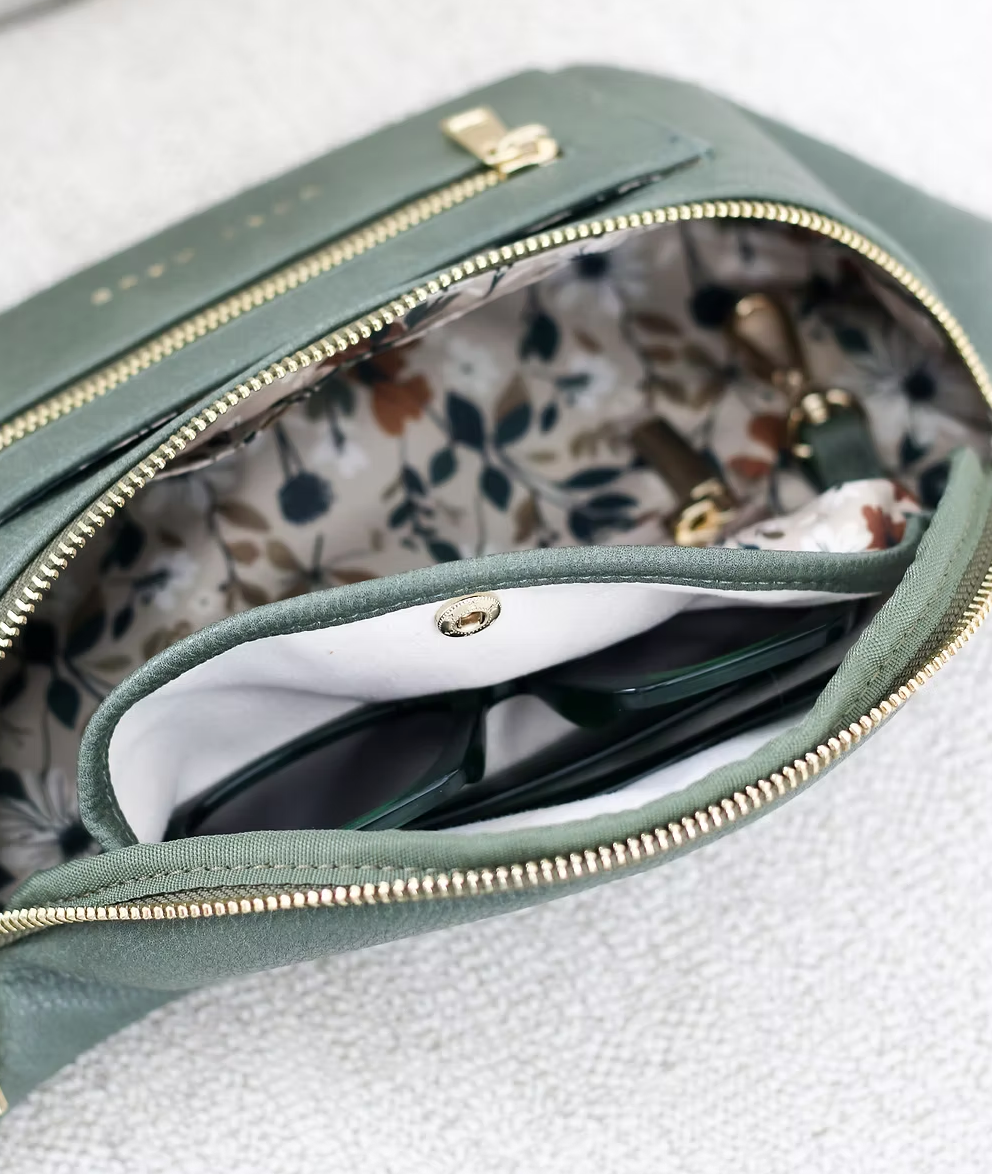 A close-up shot shows the inside of a green belt bag. The lining has a light-colored background with a pattern of brown, green, and white leaves and flowers. A pair of dark-framed sunglasses is partially visible in a separate, lighter-colored inner pocket that closes with a gold-toned snap. The main compartment has a gold zipper that is partially open.