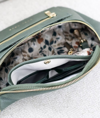 Thumbnail for A close-up shot shows the inside of a green belt bag. The lining has a light-colored background with a pattern of brown, green, and white leaves and flowers. A pair of dark-framed sunglasses is partially visible in a separate, lighter-colored inner pocket that closes with a gold-toned snap. The main compartment has a gold zipper that is partially open.