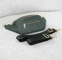 Thumbnail for A green belt bag with gold zippers and two different sized green straps are placed on a textured white surface. The words 