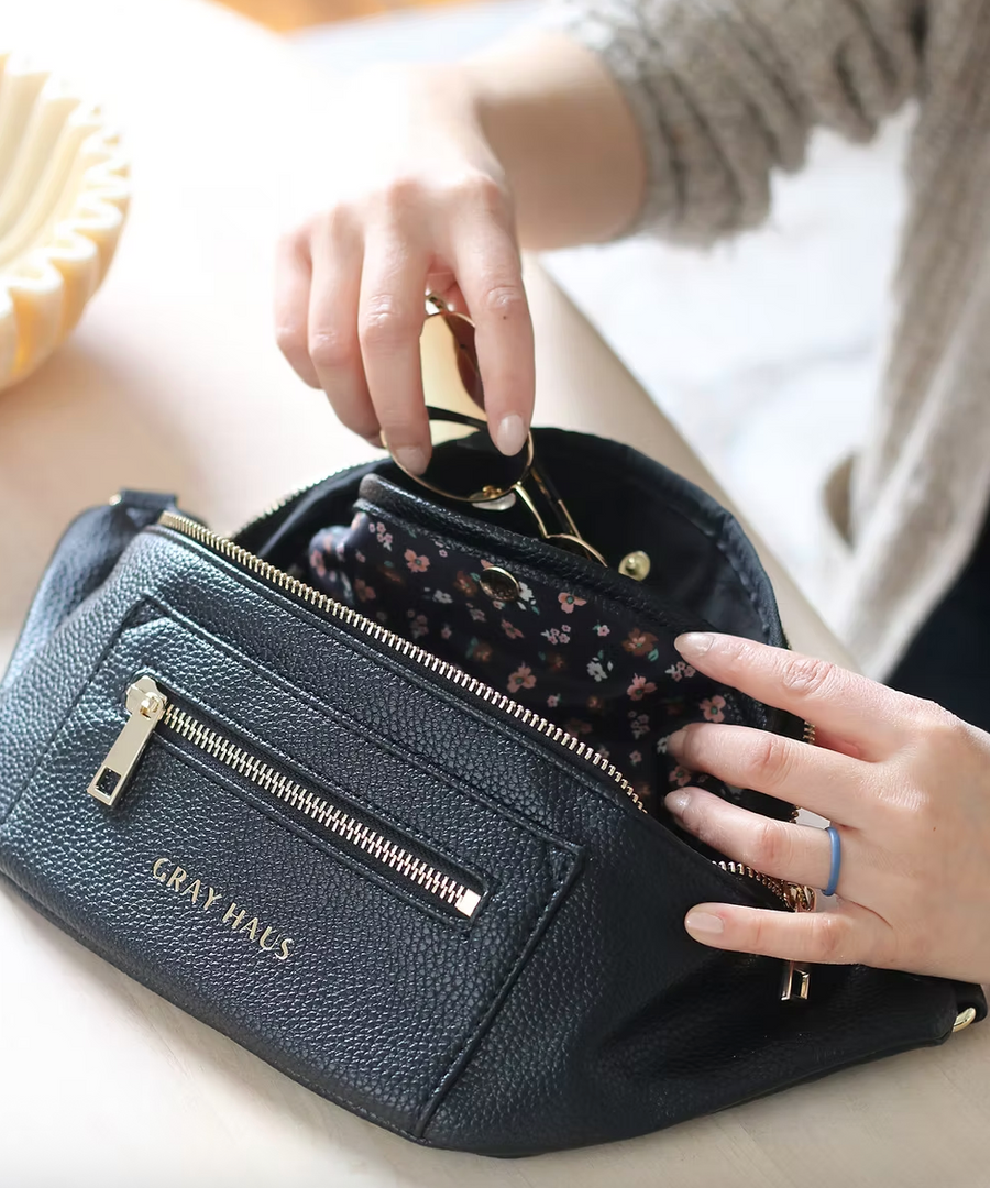 A black belt bag is partially open, revealing a floral lining. A hand is reaching into the bag. The bag features two zippered compartments on the front, and the words "GRAY HAUS" are subtly embossed below the smaller front zipper. The top zipper extends across the full width of the bag. The adjustable belt strap is partially visible.