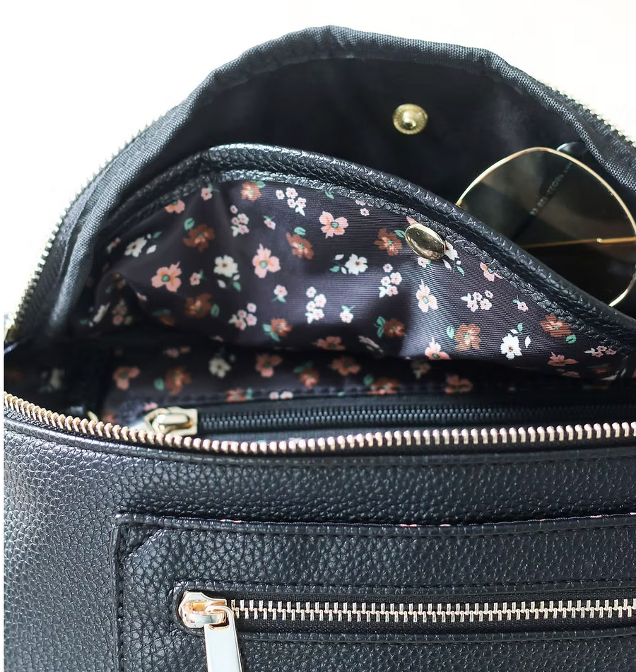 A close-up shot shows the inside of a black belt bag. The lining is a dark fabric with a pattern of small pink and white flowers. A pair of sunglasses with gold frames is partially visible inside a snap section of the main compartment. There's a smaller zippered pocket within the main compartment. The exterior of the bag, made of textured black material, features a gold zipper for the main compartment and a portion of a smaller zippered pocket below it.