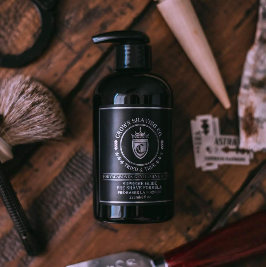 This image shows a black pump bottle of Crown Shaving Co. Supreme Glide Pre-Shave Formula. The label prominently displays the brand's logo and the product name "SUPREME GLIDE PRE SHAVE FORMULA PRÉ-RASER LA FORMULE." The bottle has a black pump dispenser and is set against a blurred background, possibly suggesting a bathroom or grooming area.