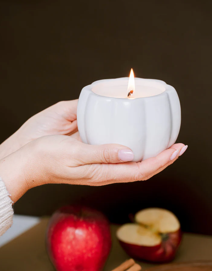 A pair of hands gently holds a white, pumpkin-shaped candle holder with a lit wick. A whole and a half-cut apple are visible in the blurred foreground.