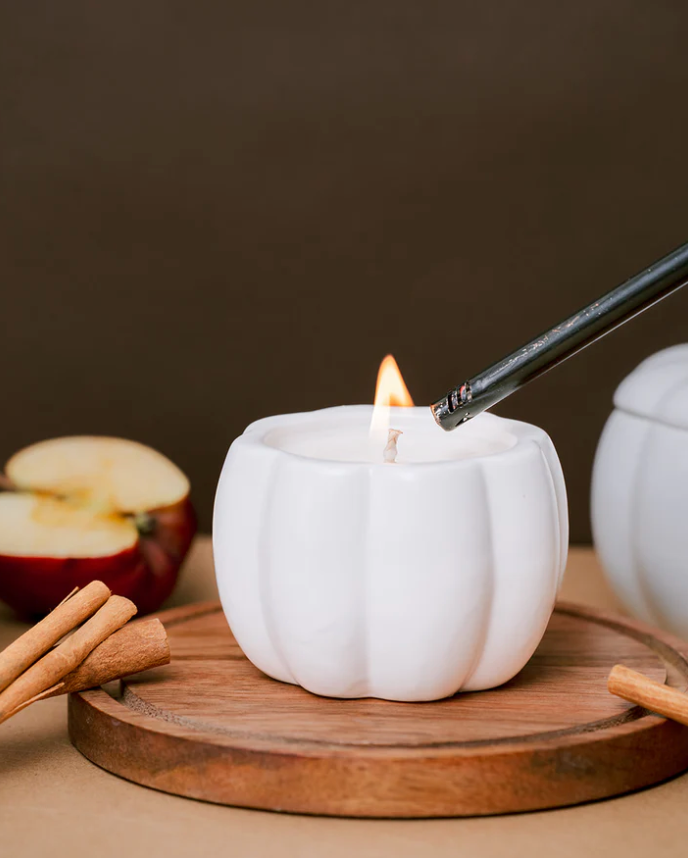 A person uses a long lighter to light the wick of a white, pumpkin-shaped candle holder. The candle is on a wooden tray with cinnamon sticks and a sliced apple.