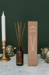 Thumbnail for 
A brown glass Boho + Glow reed diffuser with wooden reeds inserted stands next to its tall, matching brown cardboard box. A brass candle holder with a white taper candle and two metallic ornaments are visible around the diffuser, all set against a dark green background.