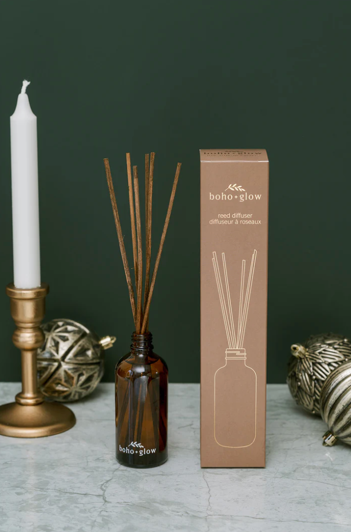 
A brown glass Boho + Glow reed diffuser with wooden reeds inserted stands next to its tall, matching brown cardboard box. A brass candle holder with a white taper candle and two metallic ornaments are visible around the diffuser, all set against a dark green background.