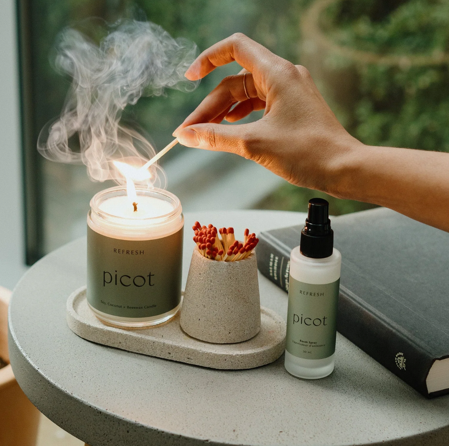 A hand holds a lit match above a smoking "picot REFRESH" candle on a concrete tray, next to a match holder filled with red-tipped matches and a "picot REFRESH" room spray bottle. A book rests in the blurred background.