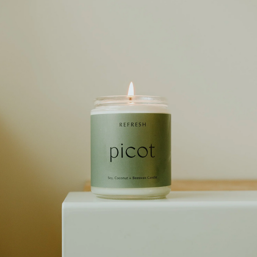 A lit jarred candle sits on a white surface against a muted beige background. The candle has a sage green label that reads "REFRESH" at the top and "picot" in a larger, lowercase, sans-serif font in the center. Below that, in smaller text, it says "Soy, Coconut + Beeswax Candle". The warm glow of the flame is visible above the white wax in the clear glass jar