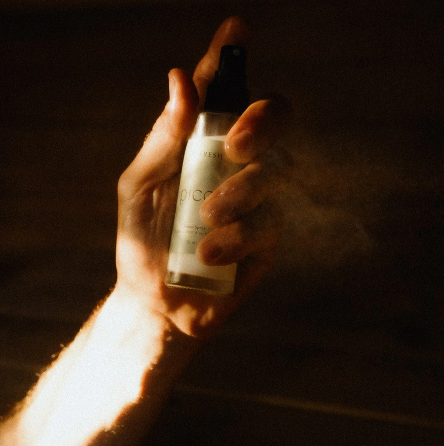 A hand holds the "picot refresh" room spray bottle, with a fine mist of spray visible coming from the nozzle. The hand is illuminated by a warm, golden light.