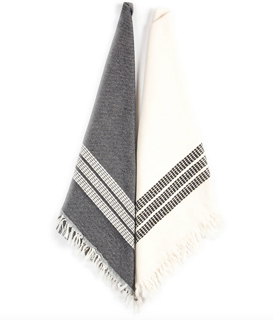 Two Rhodes Cotton Kitchen Towels are hanging side-by-side against a white background. The towel on the left is mostly dark gray with a white section near the bottom featuring horizontal bands of small black dashes and white stripes, finished with a fringed edge. The towel on the right is mostly white with a similar band of black dashes and white stripes near the bottom, also with a fringed edge.