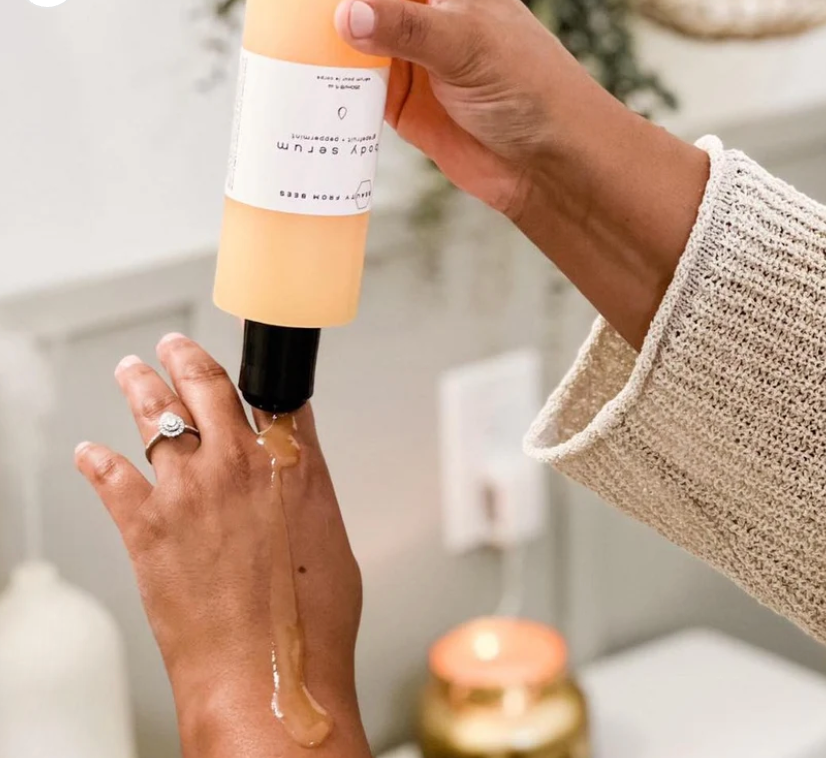 A person's hands are shown dispensing a light brown, viscous liquid labeled "body serum" from a white bottle with a black pump. The serum pours down the back of their other hand, which is adorned with a silver ring featuring a central stone.