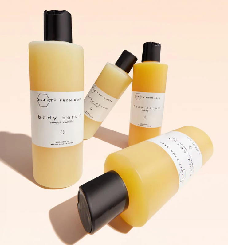 Four bottles of Beauty From Bees body serum are arranged on a light surface. Two bottles are upright, one labeled "sweet vanilla" and the other "orange". Two more bottles lie horizontally, also appearing to be different scents from the same product line. All bottles are a similar cylindrical shape with black flip-top caps and white labels featuring the brand name and scent.