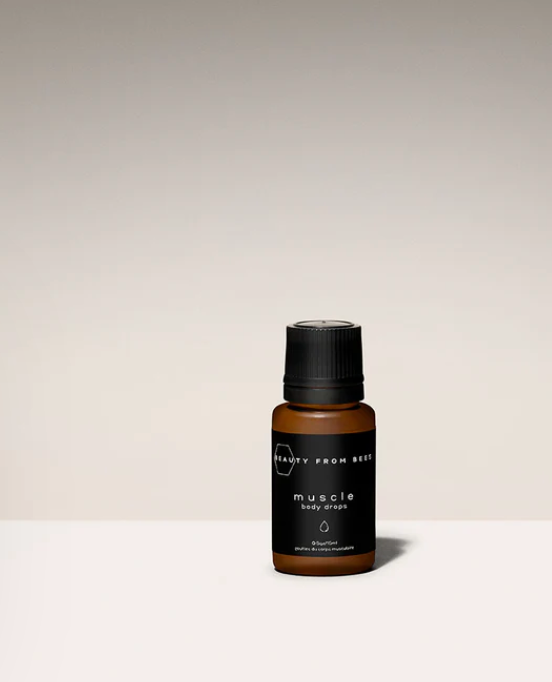 A small, dark amber glass bottle with a black ribbed cap sits centered on a light surface against a gradient beige background. The bottle has a black label that reads "BEAUTY FROM BEES" at the top and "muscle body drops" in a larger, stylized font below. A small droplet icon is also visible on the label. The bottle casts a subtle shadow to its right.