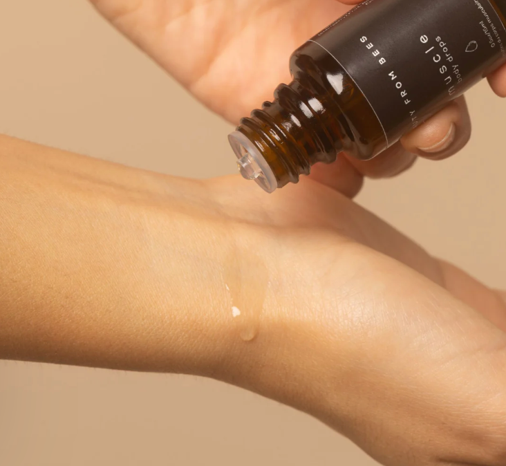 A person's hands are shown dispensing clear drops of "muscle body drops" from a small, dark amber glass bottle onto their wrist. The bottle has a black label with white text, partially visible, reading "BEAUTY FROM BEES" vertically and "muscle body drops" horizontally.