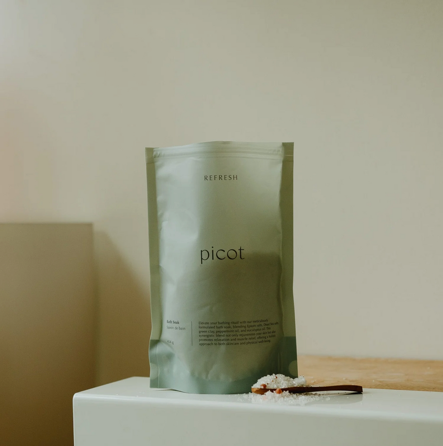 A sealed package of  bath soak, labeled 'picot', placed on a surface with a small amount of bath salts spilled on a wooden spoon in front.