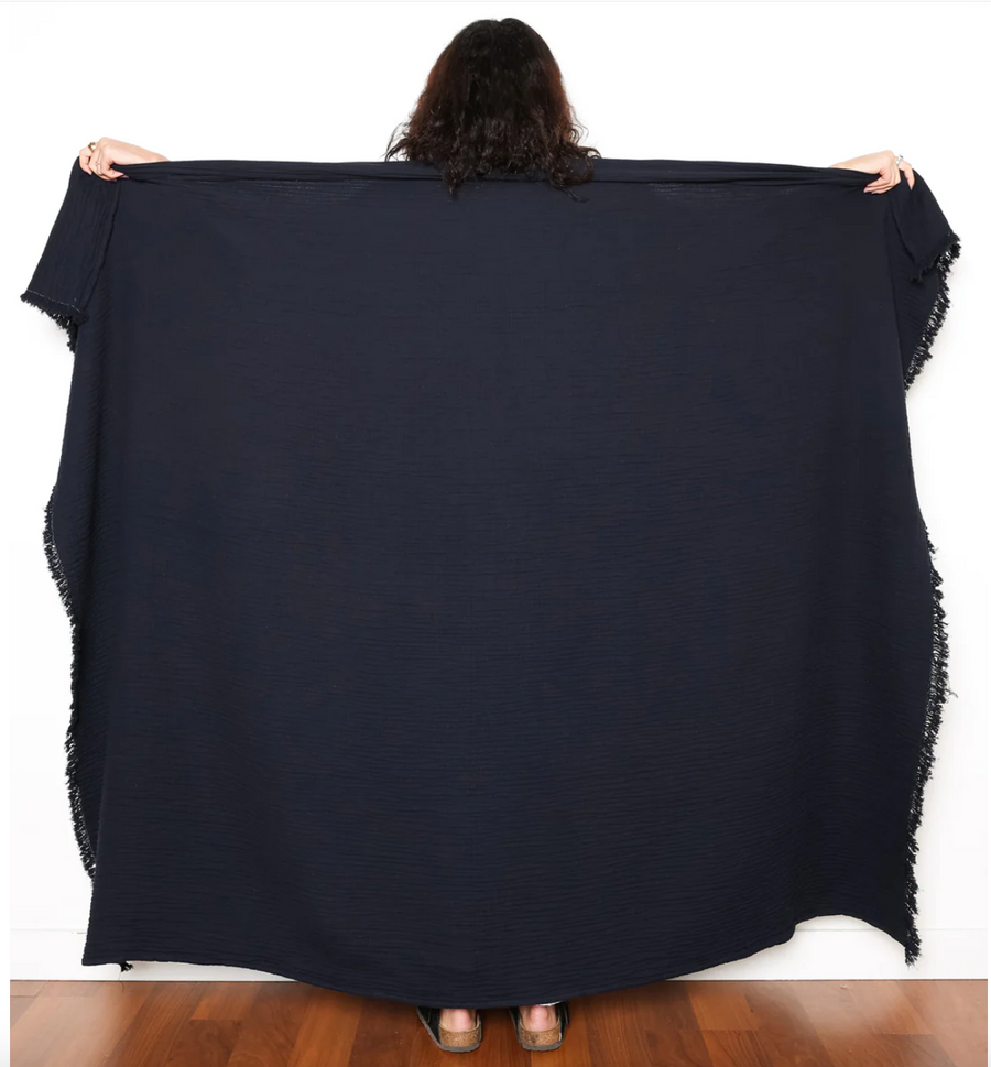 A person, viewed from the back, is holding up a large, black Capella Muslin Throw with fringed edges. The throw extends to the light wood floor and is displayed against a white wall.