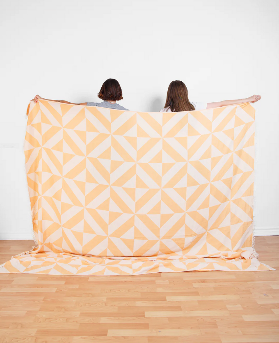 Two people, viewed from the back, are holding up a large, rectangular Como X-Large Beach Throw, which has a geometric pattern of overlapping off-white and light orange triangles. The throw extends to the light wood floor and is displayed against a white wall.