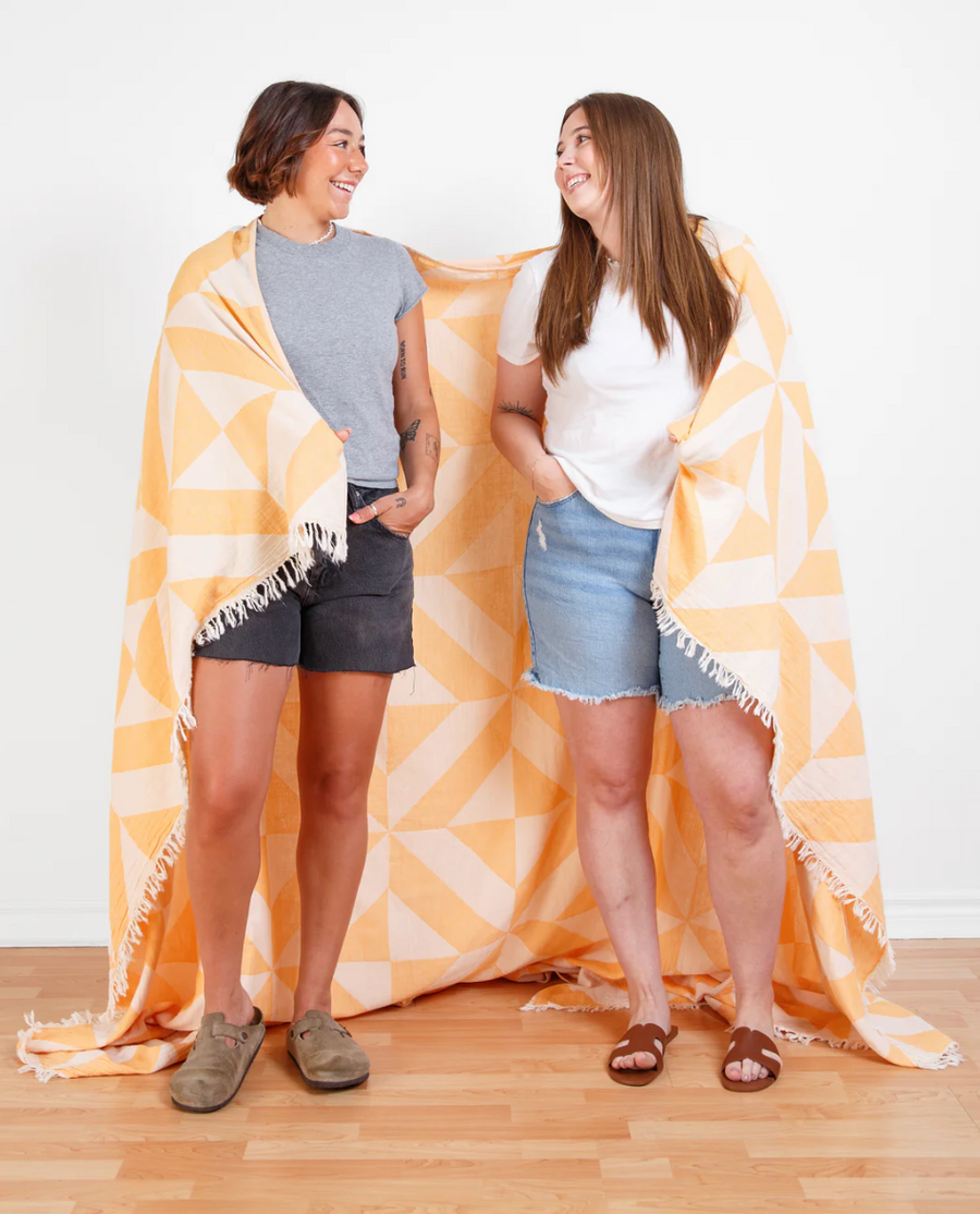 Two people are standing, wrapped in a large, rectangular Como X-Large Beach Throw, which has a geometric pattern of overlapping off-white and light orange triangles. The throw extends to the light wood floor and is displayed against a white wall.