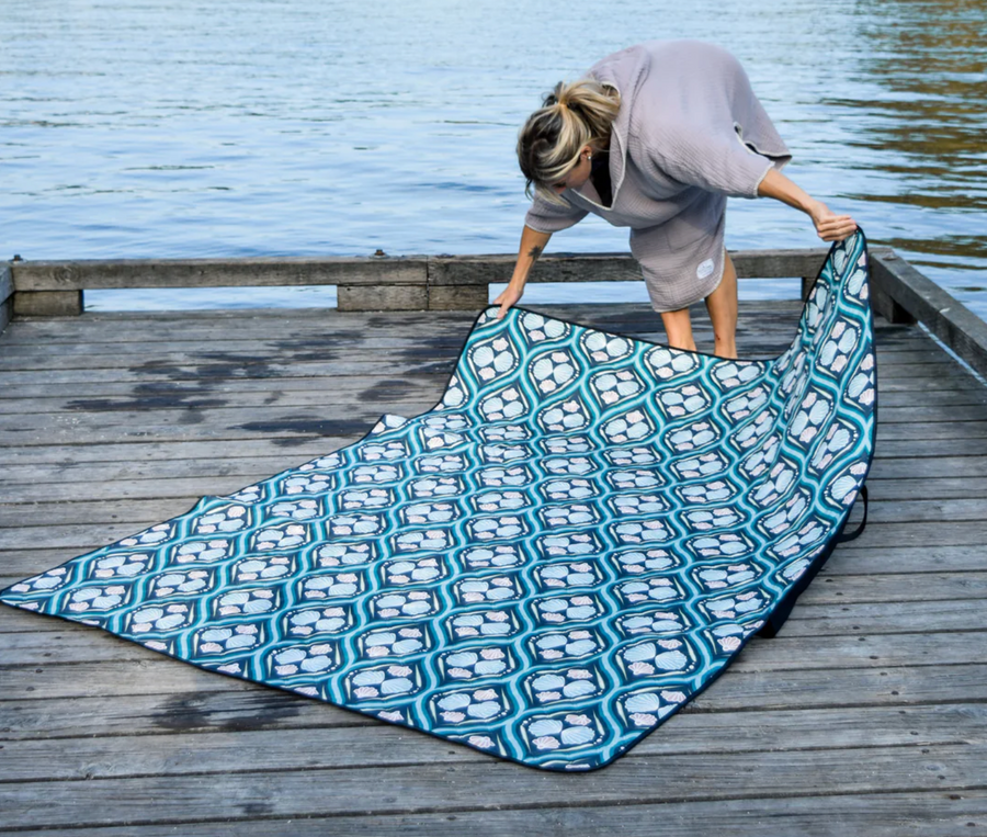 A person wearing a gray hooded towel robe is laying out a large, patterned picnic blanket on a weathered wooden dock next to a body of water. The blanket has a repeating geometric design in shades of blue and white with a black border. The person is bending over, spreading the blanket out.