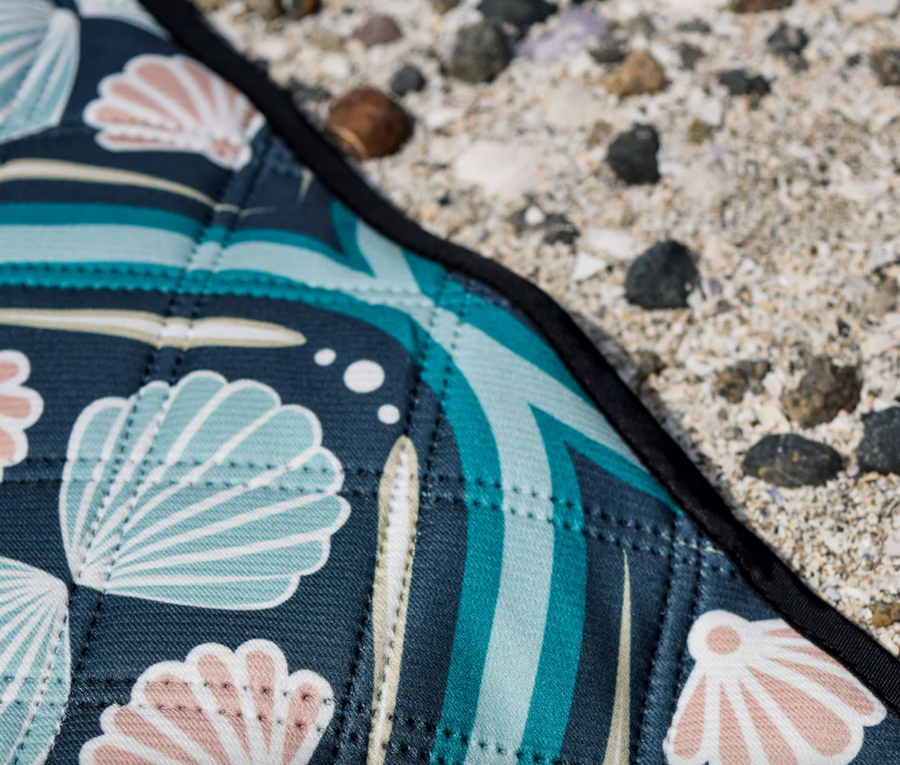 A close-up shot shows the corner of a patterned picnic blanket laid out on a surface of sand and small rocks. The blanket features a design with dark blue, teal, and light blue wavy lines, white dots, and stylized seashell shapes in shades of light pink and white. A black border runs along the edge of the blanket. The stitching of the quilted fabric is also visible.