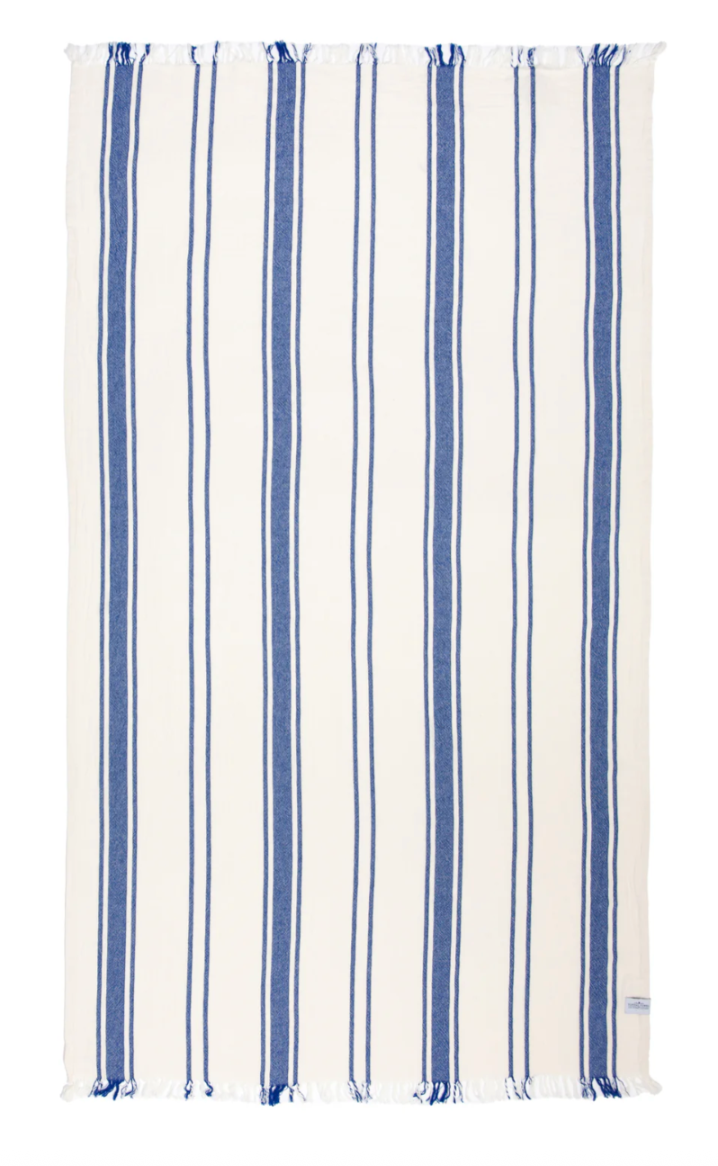 
A rectangular beach towel with a white background and evenly spaced vertical stripes in two shades of blue, alternating between a thinner and a thicker stripe. Both the top and bottom edges of the towel have white fringe with hints of blue. A small label is visible on the bottom right corner.