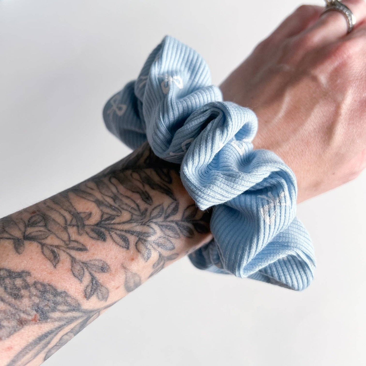 A light blue scrunchie with a ribbed texture and small white bow-tie shapes is worn on a wrist with a tattoo on the wrist/arm.