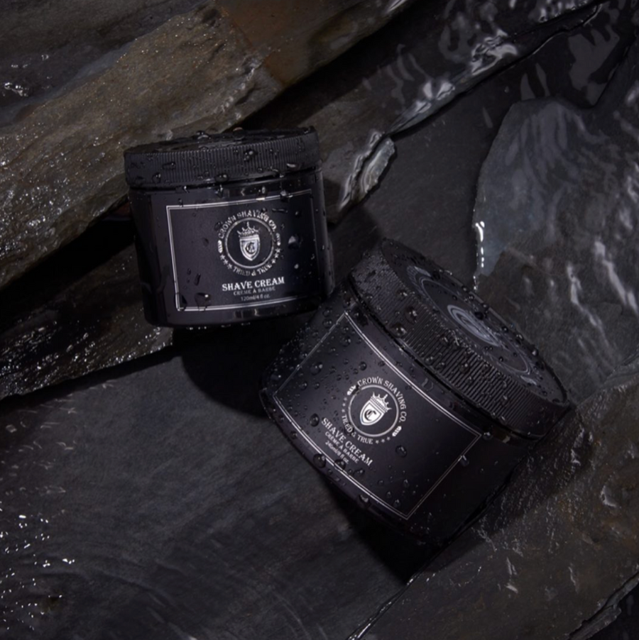 This image features two black jars of Crown Shaving Co. Shave Cream, partially submerged in water amongst dark, textured rocks. Water droplets cover the jars, and the labels with the brand's logo and product name are visible. The lighting creates a dramatic and moody atmosphere.