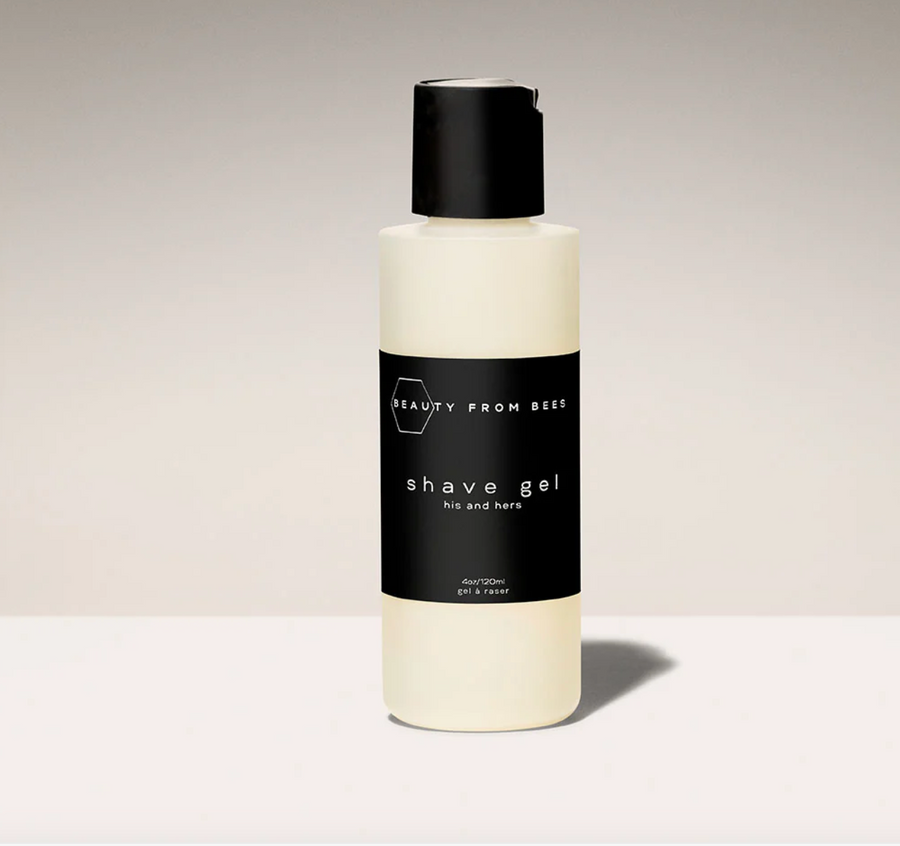 A white bottle of "BEAUTY FROM BEES" shave gel stands upright on a light surface against a pale background. The bottle has a black label with white text reading "shave gel" in a prominent font and "his and hers" in a smaller font below. A black flip-top cap is on the top of the bottle.