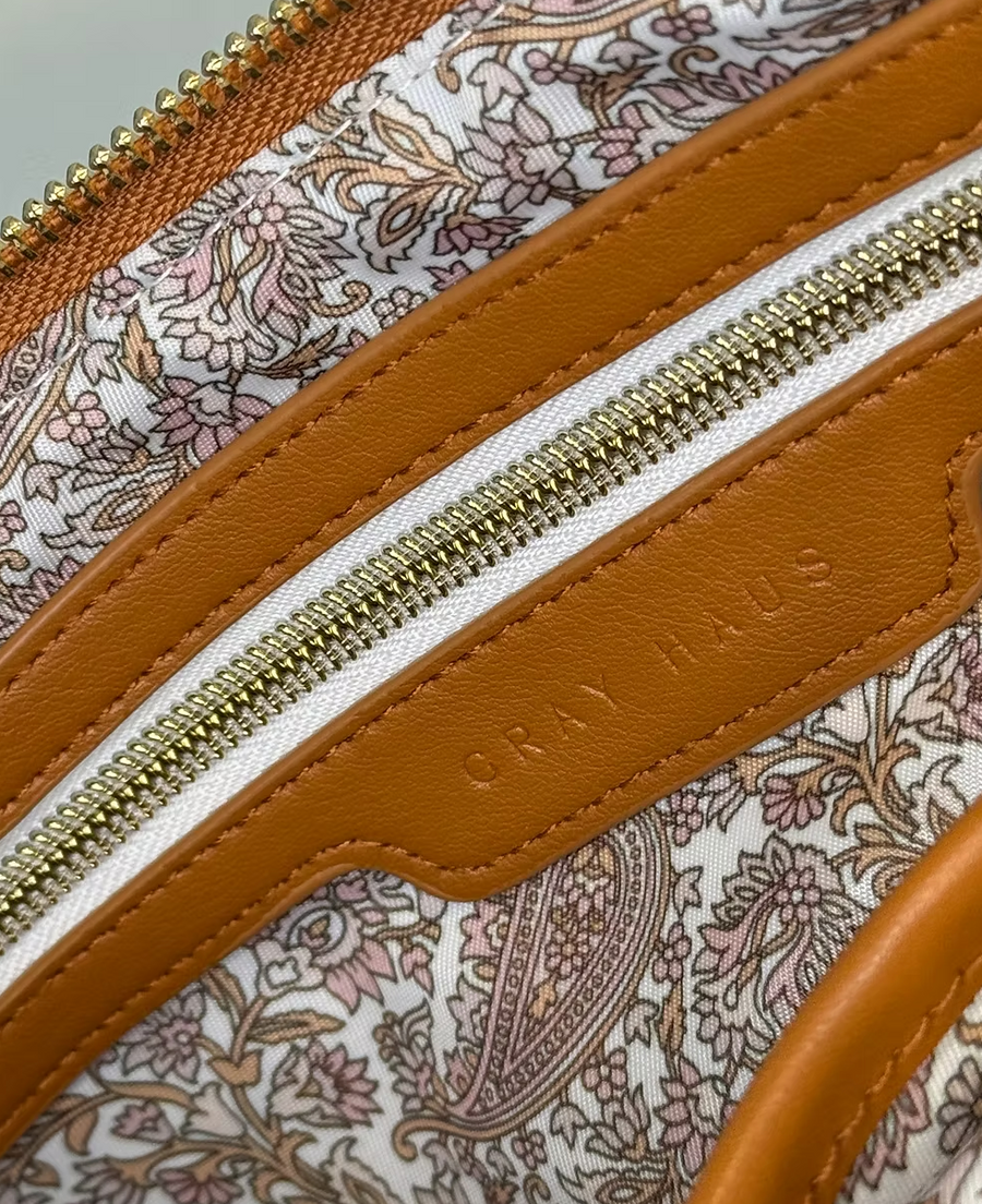 
A close-up shot of a brown crossbody bag, partially open, reveals a light-colored lining with a paisley pattern. Tan vegan leather trim and gold zippers are also visible. The words "GRAY HAUS" are visible.