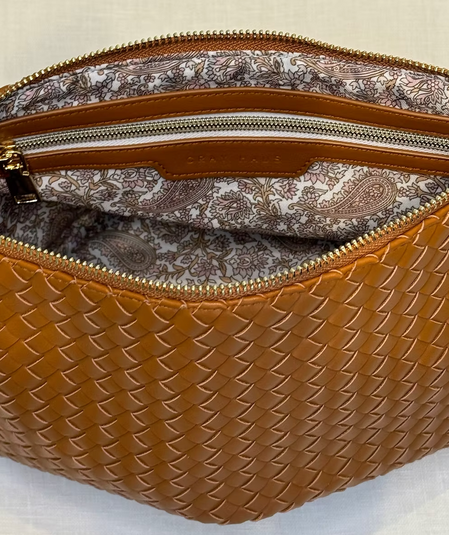 A brown, woven crossbody bag is shown with its main compartment partially open, revealing a light-colored interior lining with a paisley pattern in shades of brown and pink. A smaller zippered pocket is visible inside. The bag features gold zippers and a tan vegan leather trim along the zipper openings. The woven texture of the brown exterior is clearly visible.