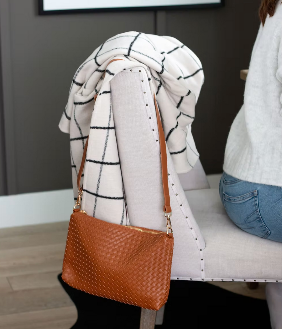 A brown woven bag with a gold zipper is hanging from the arm of a chair. A white and black plaid blanket is draped over the chair. A woman wearing jeans and a white sweater is sitting in the background.