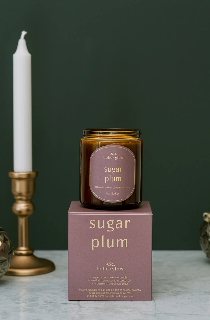 
An amber jar candle labeled "sugar plum" by Boho + Glow sits on top of its matching muted purple box. A brass candle holder with a white taper candle is partially visible on the left against a deep green background.