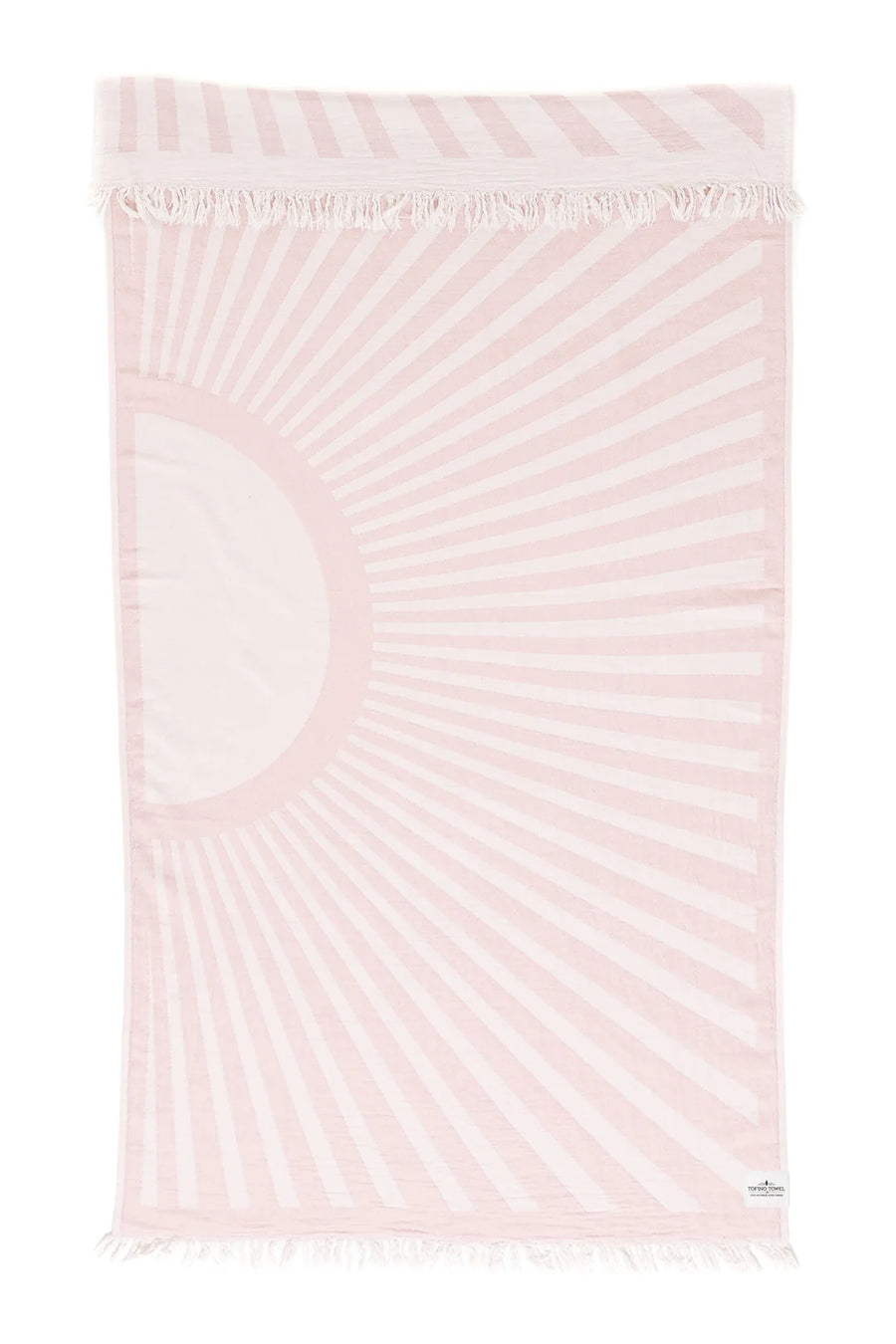 
A rectangular "Sun Flare - Rose Smoke" beach towel with a white background and a large sunburst design in light pink. The sun is on the left side, emitting rays across the towel. Both the top and bottom edges of the towel have white fringe. A small white label is visible on the bottom right corner.