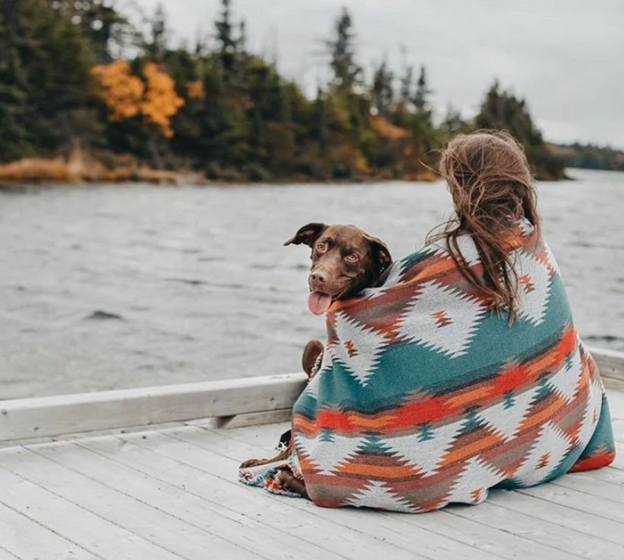 
A woman with long brown hair, seen from behind, sits on a wooden dock wrapped in a Southwestern-patterned blanket in shades of teal, orange, white, and brown. A brown dog is nestled beside her, looking over her shoulder towards the viewer with its tongue slightly out. In the background, a calm body of water stretches out to a tree-lined shore with hints of autumn foliage. The sky is overcast.