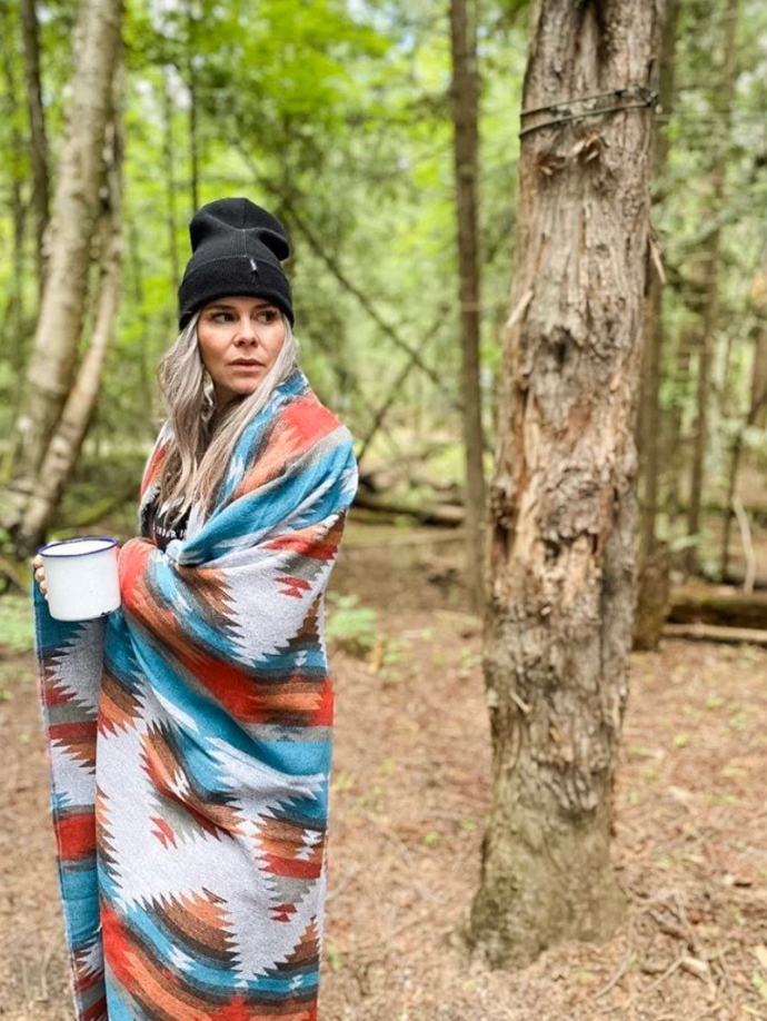 
A woman with long, light-colored hair and a black beanie stands in a forest, wrapped in a Southwestern-patterned blanket of teal, orange, white, and brown. She holds a white mug in her left hand and looks towards the left side of the frame with a thoughtful expression. Tall trees with green foliage surround her.