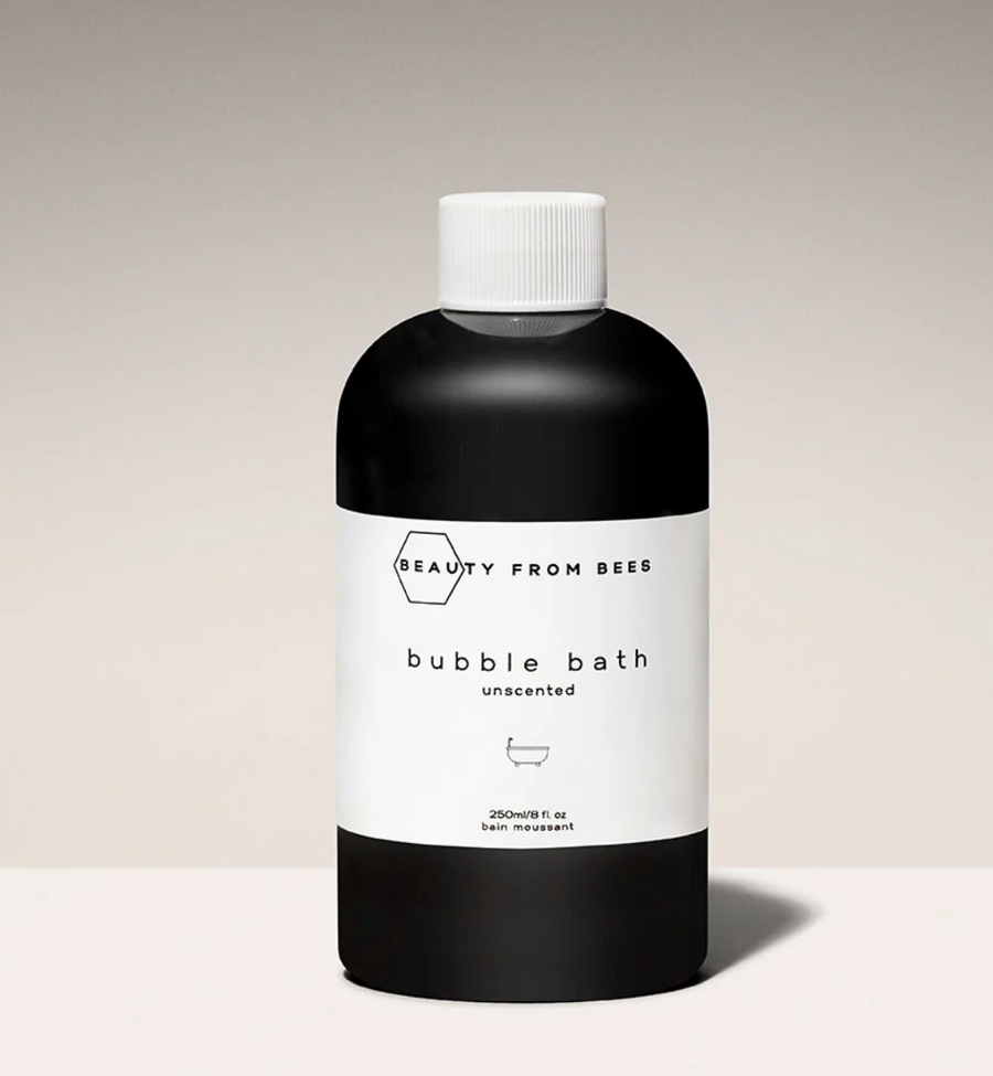 A black bottle of "BEAUTY FROM BEES" bubble bath, labeled "unscented," stands centered against a gradient beige background. The bottle has a white label with black text and a simple bathtub icon. The cap of the bottle is white.