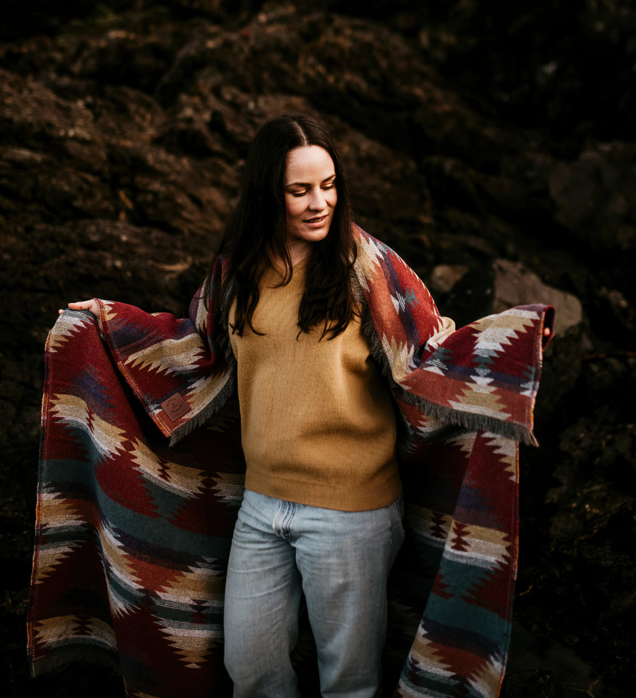A woman with long, dark hair, wearing a mustard yellow sweater and light-wash jeans, stands outdoors. She is wrapped in a thick, patterned blanket featuring geometric designs in shades of deep red, teal, orange, and beige. She looks down and to her right with a slight smile. The background is a dark, rocky terrain.