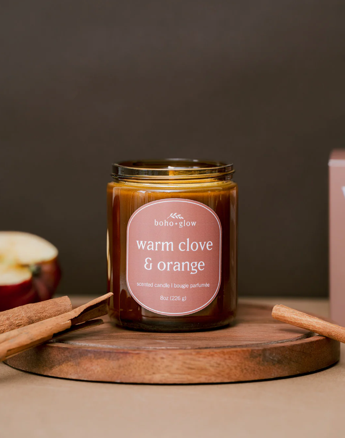 A warm clove and orange scented candle by "boho+glow" sits on a wooden board with cinnamon sticks and a cut apple.