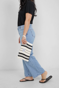 Thumbnail for A woman stands in profile wearing a black t-shirt and light-wash wide-leg jeans, carrying the ecru and black striped Mercer bag by its wrist strap at her side.