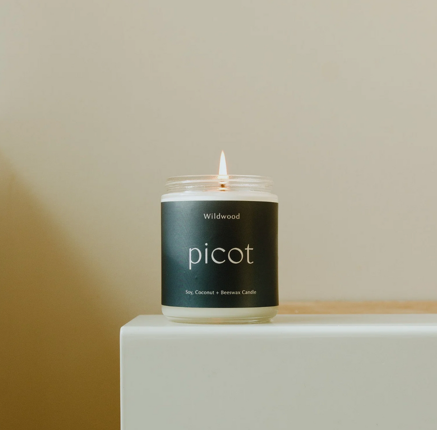 A lit jarred candle with a dark label rests on a white surface against a light beige background. The label reads "Wildwood" at the top and "picot" in a larger, lowercase, sans-serif font in the center. Below, it indicates "Soy, Coconut + Beeswax Candle". The candle's flame flickers above the white wax within the clear glass jar.